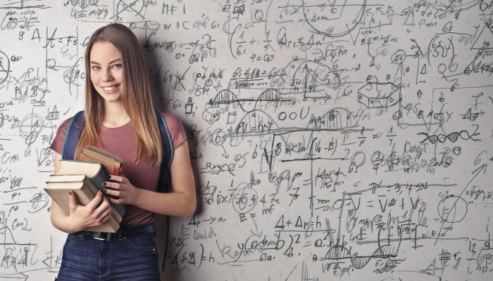 Jak skutecznie uczyć się samodzielnie? - dziewczyna z ksiązkami stoi na tle szkolnej białej tablicy z wzorami matmatycznymi